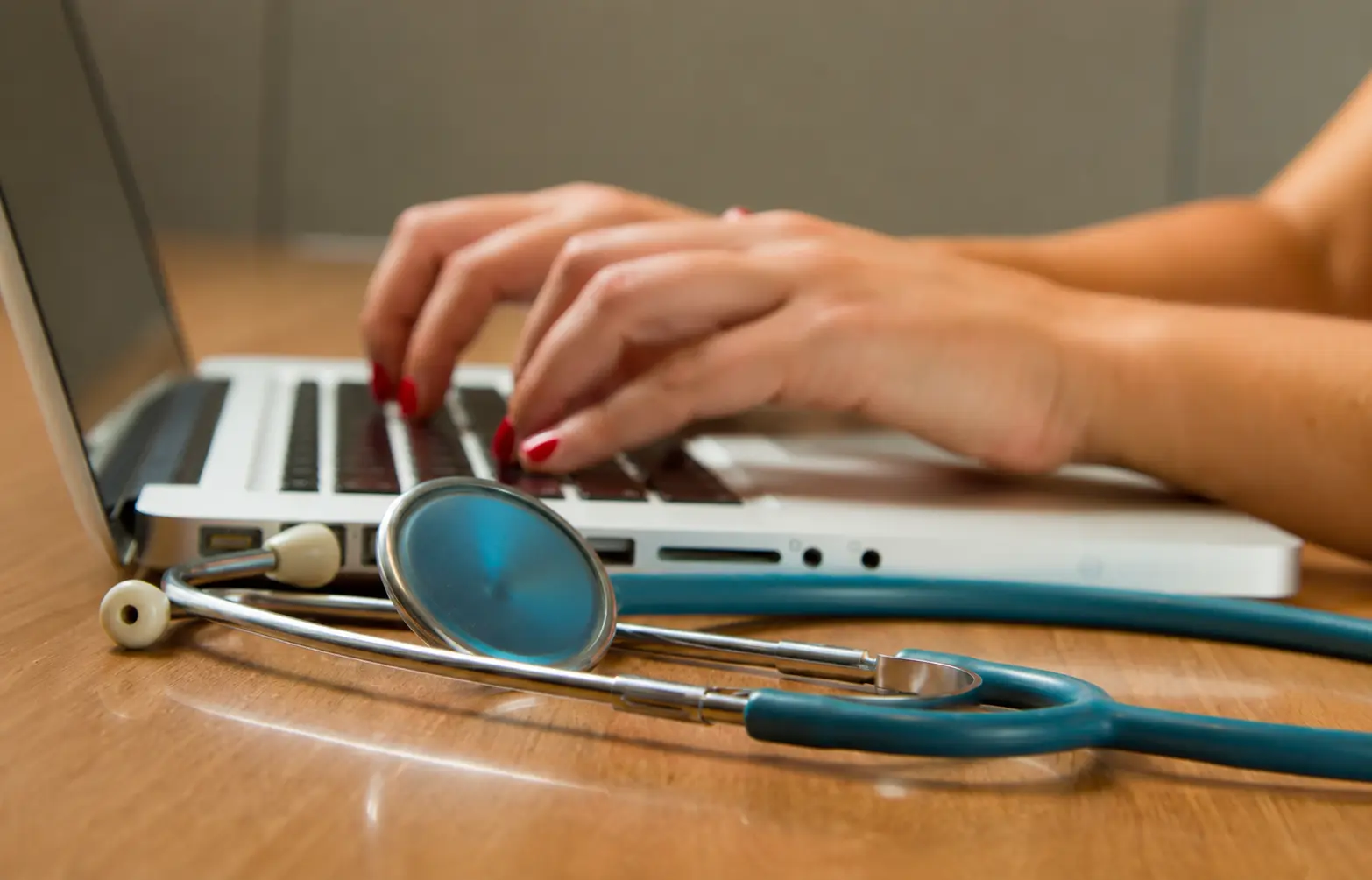 Close up of a female doctor's hands typing on a modern silver laptop on a wooden desk. A teal medical stethoscope rests in the foreground out of focus. The scene suggests meticulous medical documentation, professional evaluation, and high-quality patient care in a sophisticated clinical setting.